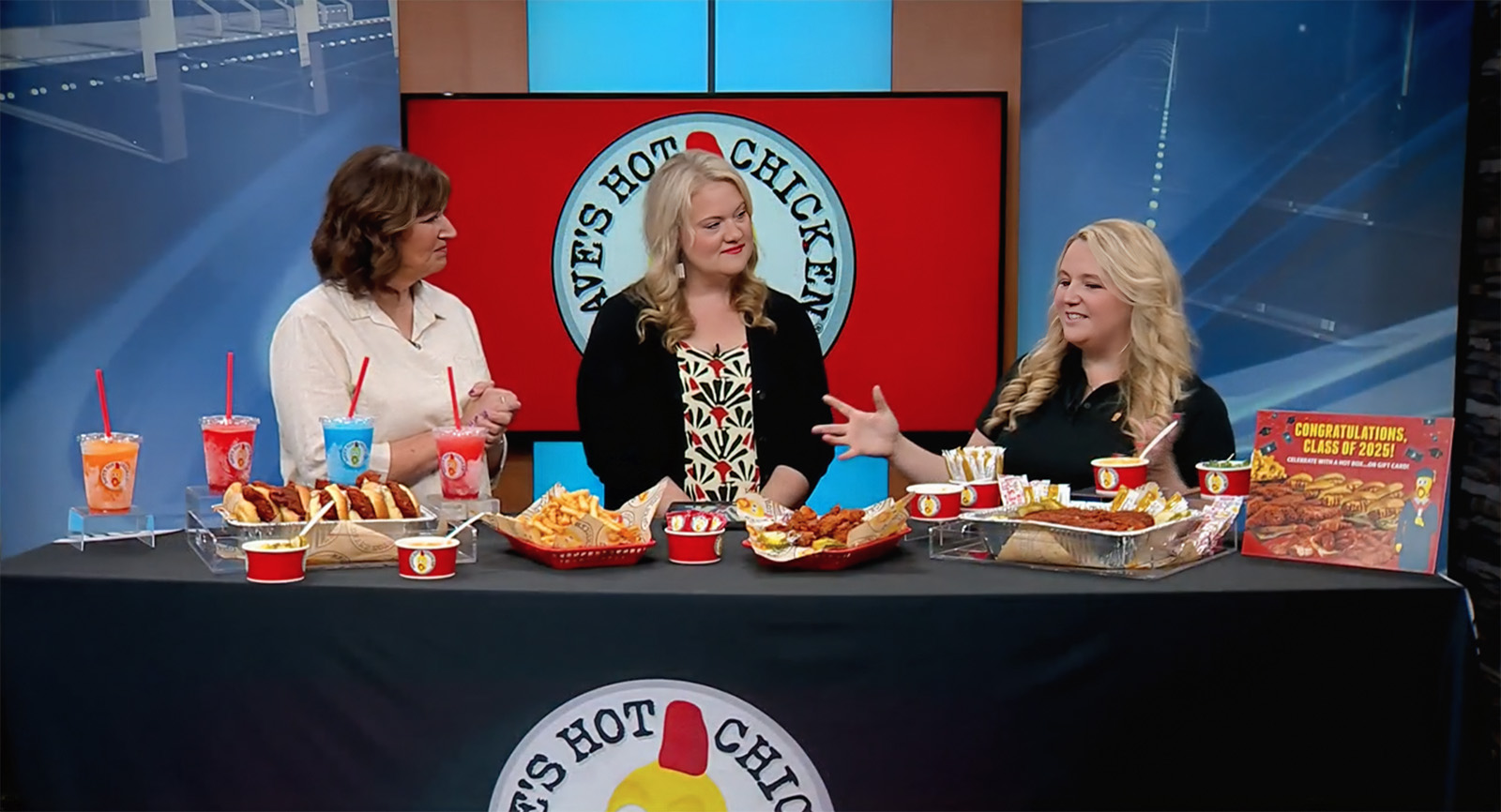 Two hosts of the morning news show Local 5 Live in Green Bay, WI, discussing Dave's Hot Chicken with a brand representative who is gesturing to the table of food in the foreground.