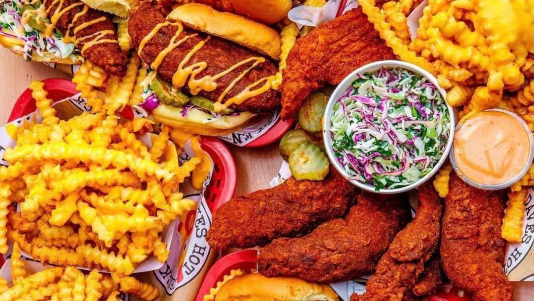 Overhead view of a Dave's Hot Chicken feast featuring Nashville-style hot chicken tenders and sliders, crinkle-cut fries topped with Dave's sauce and cheese, creamy kale slaw, pickles, and dipping sauce spread across a table.