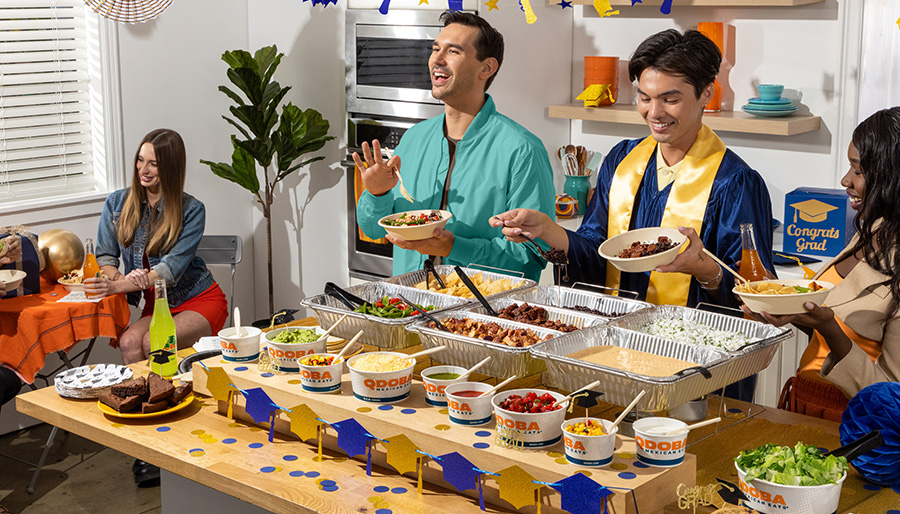 Guests gather around a QDOBA Catering hot bar during a graduation party, building customizable bowls and meals while celebrating a graduate in cap and gown.