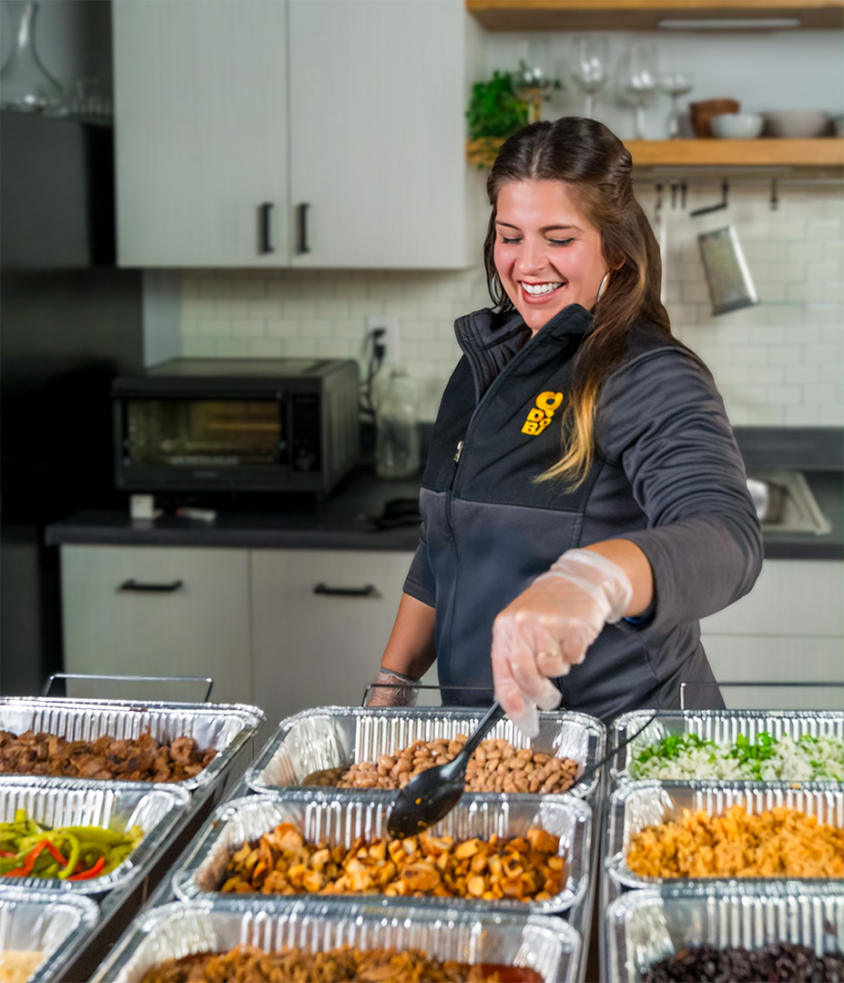 A smiling QDOBA Catering delivery driver setting up a custom Hot Bar in a client's kitchen, including hot pans of chicken, steak, rice, beans, fajitas veggies, and creamy queso, so everything will be perfectly ready for the client's guests to make their own meals.