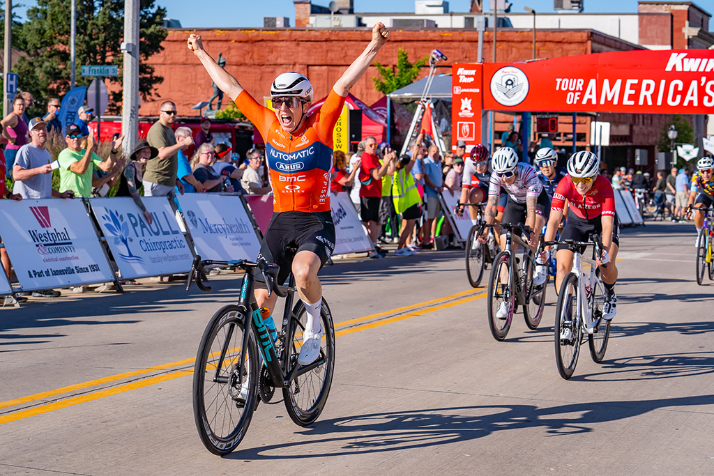Cyclists crossing the finish line. At the front, the winning cyclist wears an orange shirt and is raising his arms in celebration. In the background, spectators cheer.