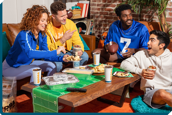 Joyful friends enjoying a QDOBA Mexican Eats catered watch party with burritos and chips, cheering on their favorite teams, showcasing a spirited home gathering with football-themed excitement and delicious food.