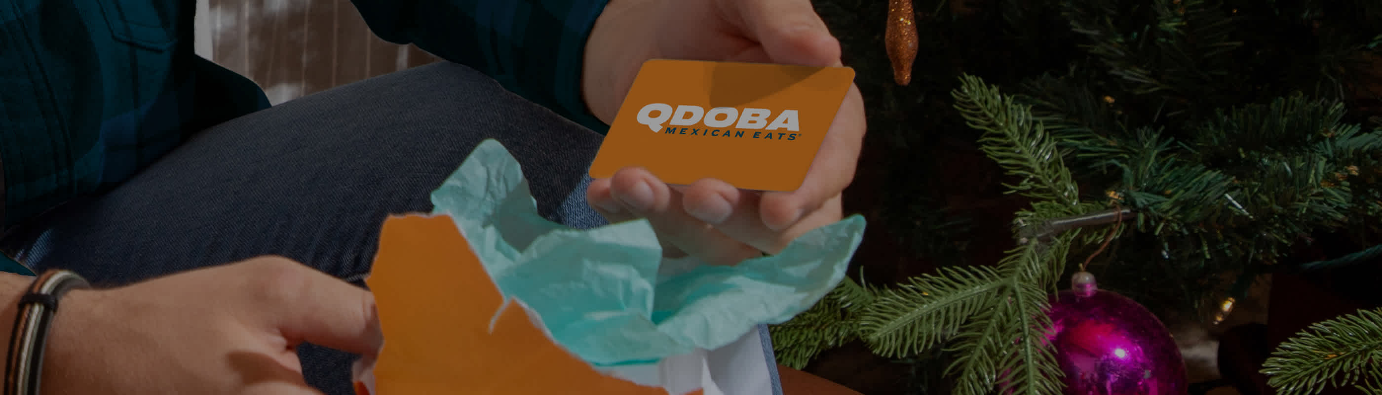 Person opening a holiday gift and holding a QDOBA Mexican Eats gift card beside a decorated Christmas tree.