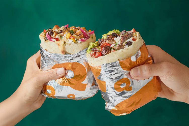Two QDOBA burritos held up in a celebratory toast, filled with fresh ingredients, promoting QDOBA West Des Moines grand opening