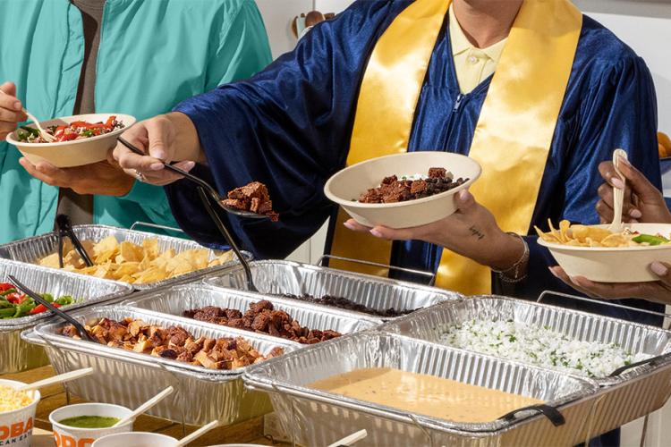 Graduation party guests build customizable QDOBA meals from a catering hot bar while celebrating a graduate in cap and gown at a graduation open house.