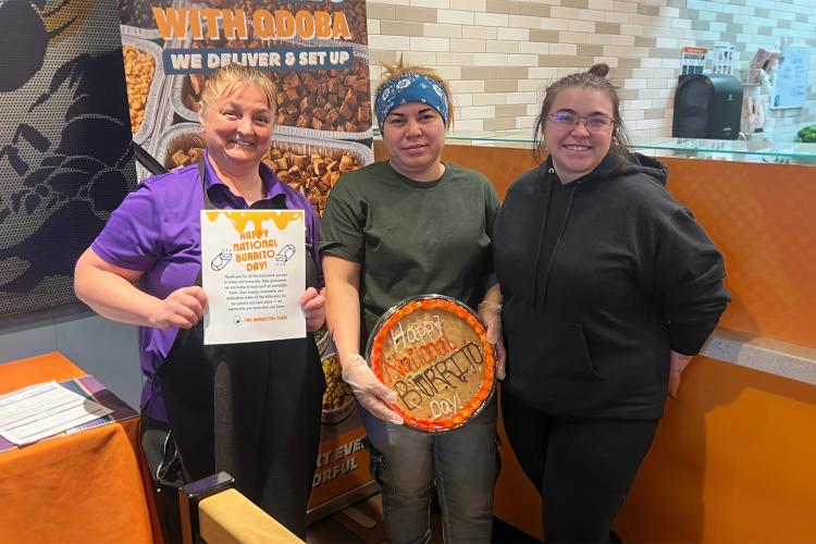 Three QDOBA team members smiling in a restaurant located in Fond du Lac, WI, holding a burrito-themed cookie cake and a “Happy National Burrito Day” appreciation sign.