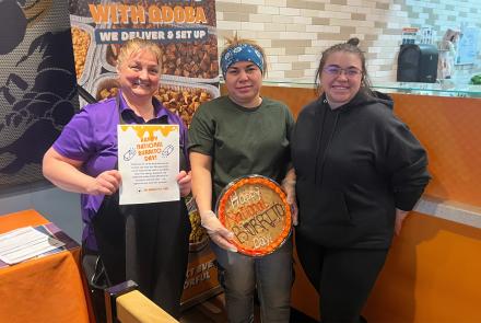 Three QDOBA team members smiling in a restaurant, holding a burrito-themed cookie cake and a “Happy National Burrito Day” appreciation sign.