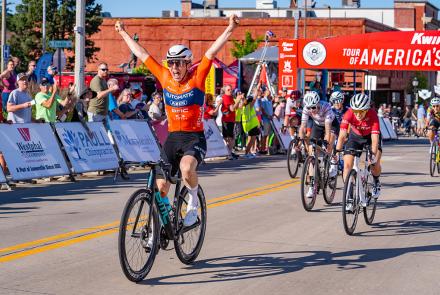 Cyclists crossing the finish line. At the front, the winning cyclist wears an orange shirt and is raising his arms in celebration. In the background, spectators cheer.