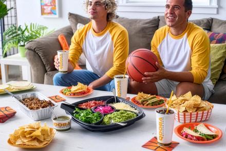 Two young men watching college basketball with a QDOBA Catering taco kit during a March Madness watch party.